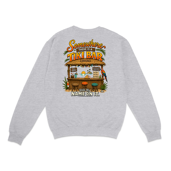 A light heather gray crewneck sweatshirt shown from the back, featuring a colorful graphic print of a tropical tiki bar with parrots and the text 'Somewhere there is a TIKI BAR with my NAME ON IT.'.