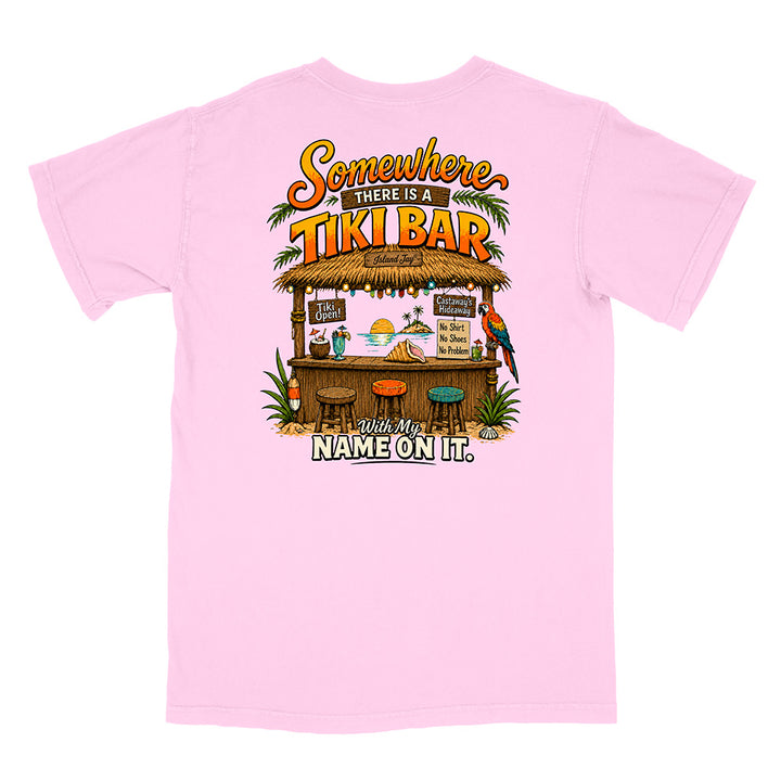 A light pink short-sleeve t-shirt with a colorful graphic on the back featuring a tiki bar illustration and the text 'Somewhere THERE IS A TIKI BAR With My NAME ON IT.'.