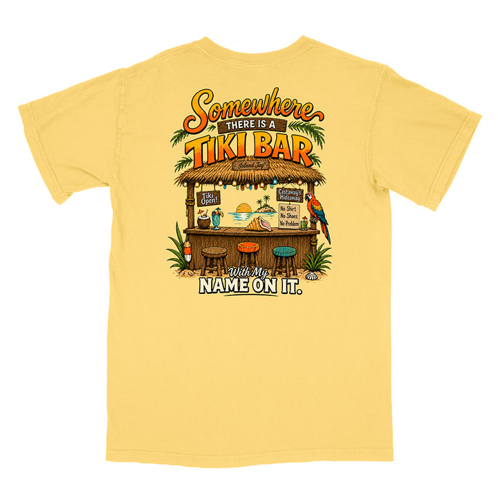 A pale yellow short-sleeve t-shirt with a large graphic print on the back depicting a tropical tiki bar scene with the text 'Somewhere THERE IS A TIKI BAR' and 'WHO'S NAME ON IT.'.