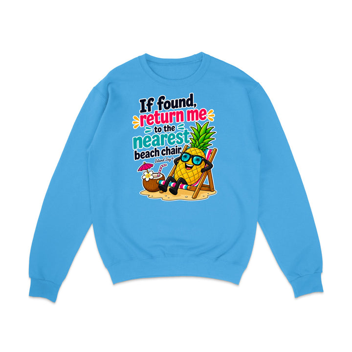 A light blue crewneck sweatshirt featuring a colorful graphic print on the front. The graphic shows a pineapple character wearing sunglasses and reclining in a beach chair on a sandy beach, with the text 'If found, return me to the nearest beach chair' in playful black, pink, orange, and teal lettering.