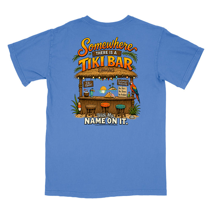 A blue short-sleeve t-shirt shown flat against a white background, featuring a large graphic print on the back with a tropical tiki bar illustration and the text 'Somewhere THERE IS A TIKI BAR With My NAME ON IT.'.