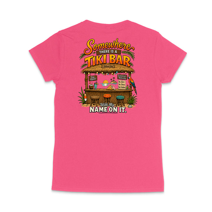 A bright pink women's short-sleeve t-shirt shown from the back, featuring a colorful graphic of a tropical tiki bar with the text 'Somewhere THERE IS A TIKI BAR' and 'With My NAME ON IT.'.