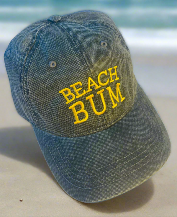 Blue denim cap with 'BEACH BUM' in yellow on a beach setting