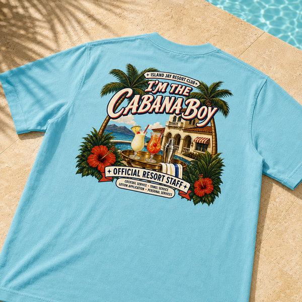 A light blue short-sleeve t-shirt laid flat on a beige stone surface near a swimming pool, showing a large colorful back graphic with palm trees, cocktails, a resort building, and the text 'I'M THE CABANA-BOY' and 'OFFICIAL RESORT STAFF'.