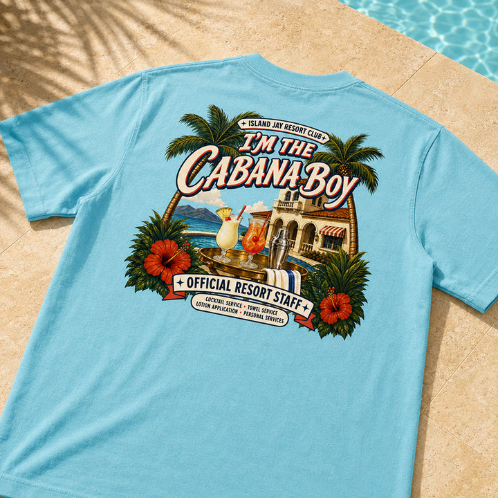 A light blue short-sleeve t-shirt laid flat on a beige stone surface near a swimming pool, showing a large colorful back graphic with palm trees, cocktails, a resort building, and the text 'I'M THE CABANA-BOY' and 'OFFICIAL RESORT STAFF'.