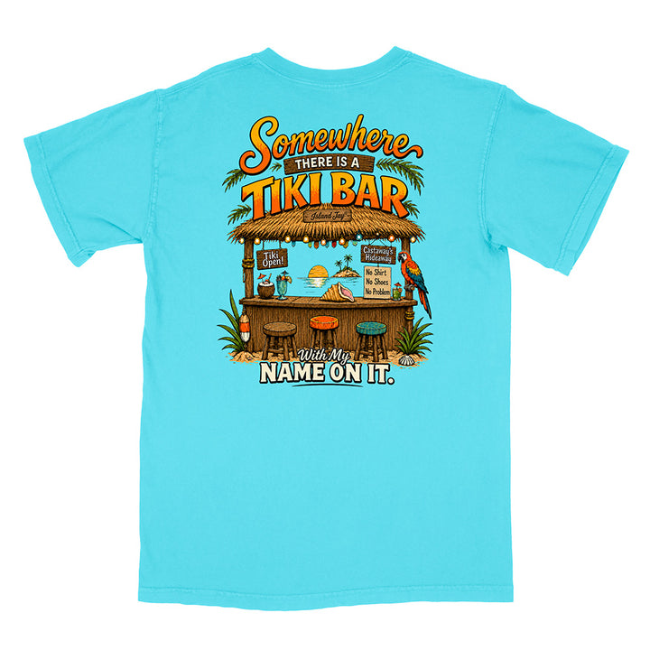 A light blue short-sleeve t-shirt with a large graphic print on the back featuring a tropical tiki bar scene and the text 'Somewhere THERE IS A TIKI BAR and my NAME ON IT.'.