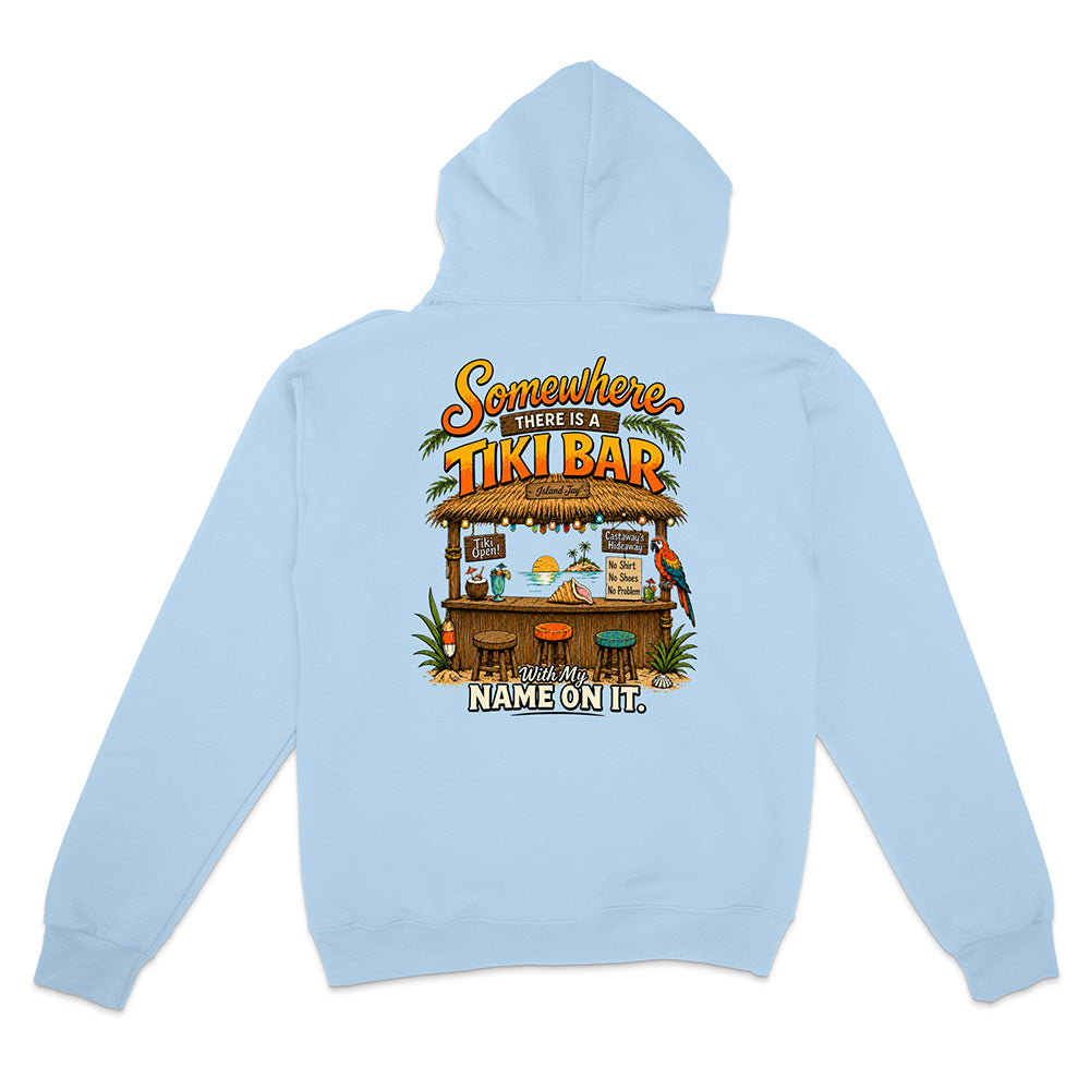 A light blue hoodie shown from the back, featuring a colorful graphic print of a tropical tiki bar with the text 'Somewhere in between a TIKI BAR' and 'NAME ON IT.'.