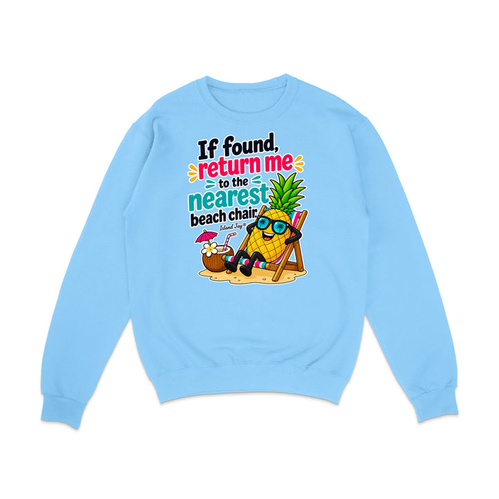 A light blue crewneck sweatshirt featuring a colorful graphic print of a pineapple character wearing sunglasses and reclining on a beach chair on a sandy beach, with the text 'If found, return me to the nearest beach chair' in stylized fonts.