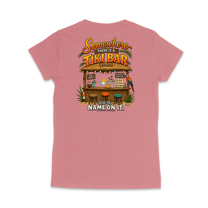 A pink short-sleeve t-shirt shown from the back, featuring a colorful graphic of a tropical 'Tiki Bar' with the text 'Somewhere there is a TIKI BAR' and 'With my NAME ON IT.'.