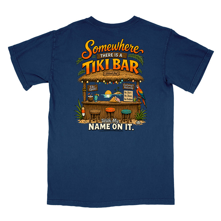 A navy blue short-sleeve t-shirt shown from the back, featuring a colorful graphic print of a tropical tiki bar with the text 'Somewhere there is a TIKI BAR' and 'Leave My NAME ON IT.'.
