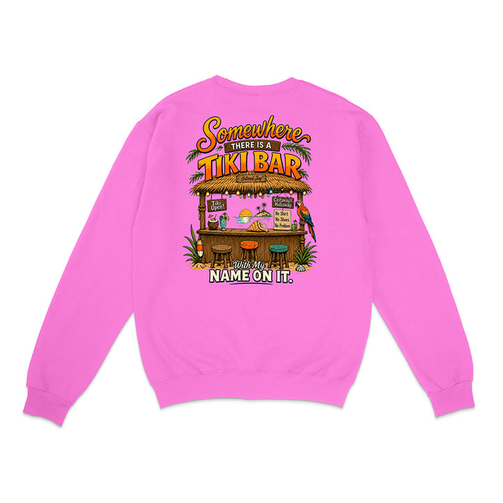 A bright pink crewneck sweatshirt shown from the back, featuring a colorful graphic of a tropical tiki bar with the text 'Somewhere there is a TIKI BAR and my NAME ON IT.'.