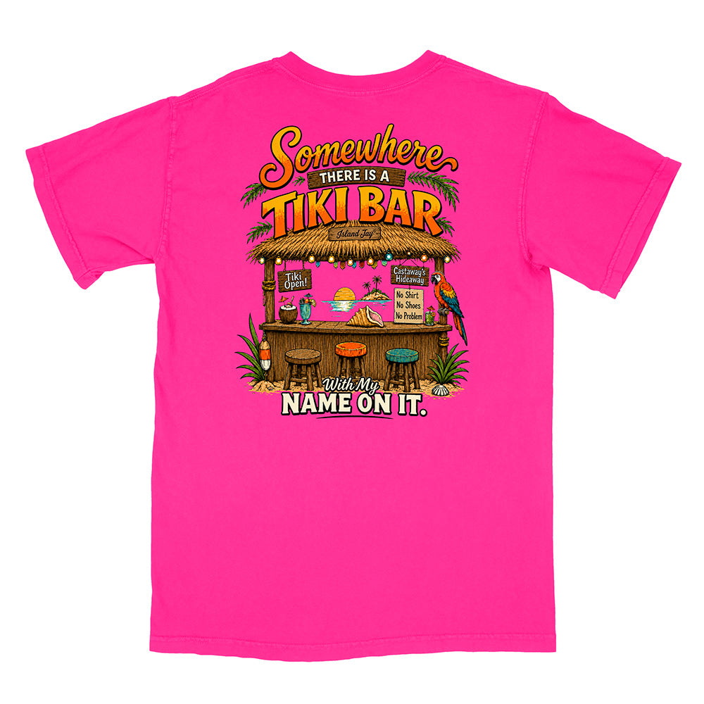A bright pink short-sleeve t-shirt with a colorful graphic on the front depicting a tropical tiki bar and the text 'Somewhere THERE IS A TIKI BAR with My NAME ON IT.'.