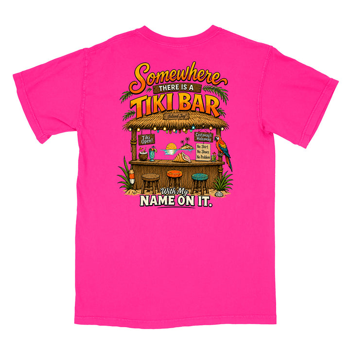 A bright pink short-sleeve t-shirt with a colorful graphic on the front depicting a tropical tiki bar and the text 'Somewhere THERE IS A TIKI BAR with My NAME ON IT.'.
