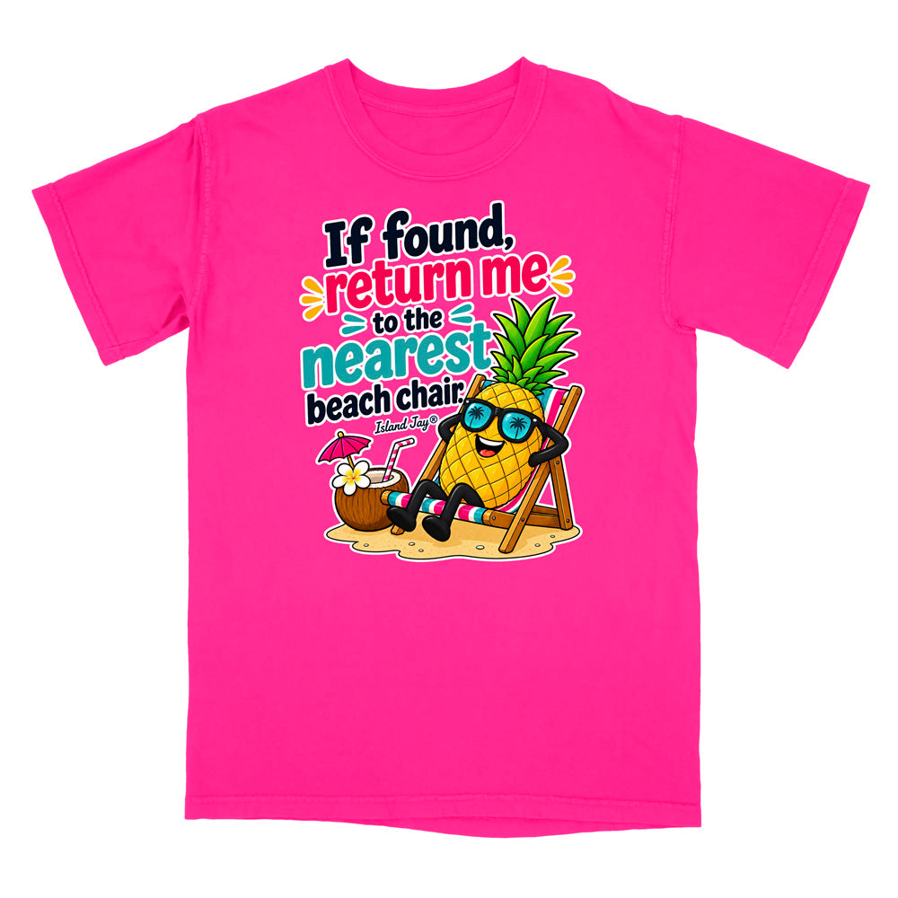 A bright pink short-sleeve t-shirt with a colorful graphic on the front. The graphic features a cartoon pineapple wearing sunglasses and reclining on a beach chair next to a coconut drink, with the text 'If found, return me to the nearest beach chair' in playful multicolor lettering.