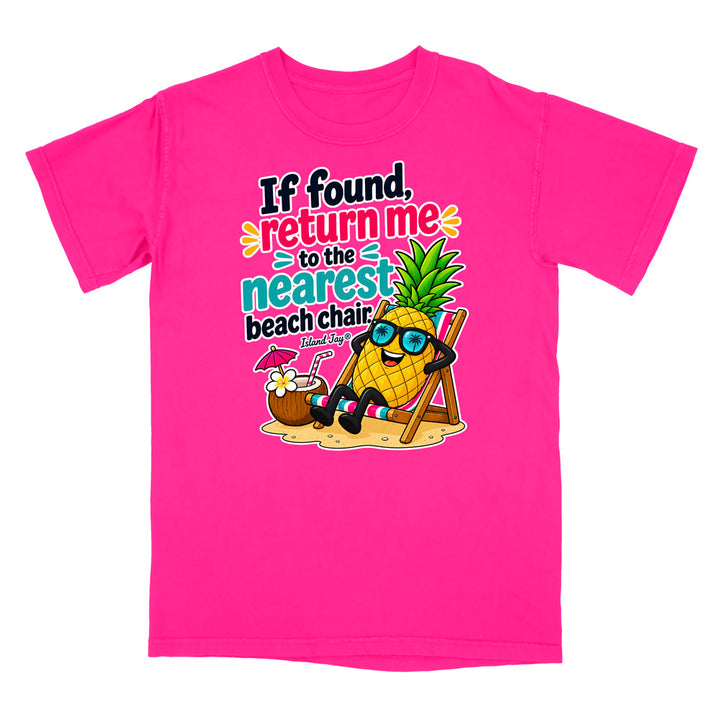 A bright pink short-sleeve t-shirt with a colorful graphic on the front. The graphic features a cartoon pineapple wearing sunglasses and reclining on a beach chair next to a coconut drink, with the text 'If found, return me to the nearest beach chair' in playful multicolor lettering.