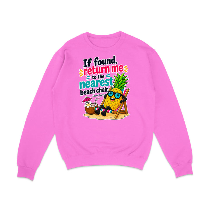 A bright pink crewneck sweatshirt featuring a colorful graphic print on the front. The graphic shows a cartoon pineapple wearing sunglasses and reclining in a beach chair next to a coconut drink, with the text 'If found, return me to the nearest beach chair' in black, teal, and orange lettering.