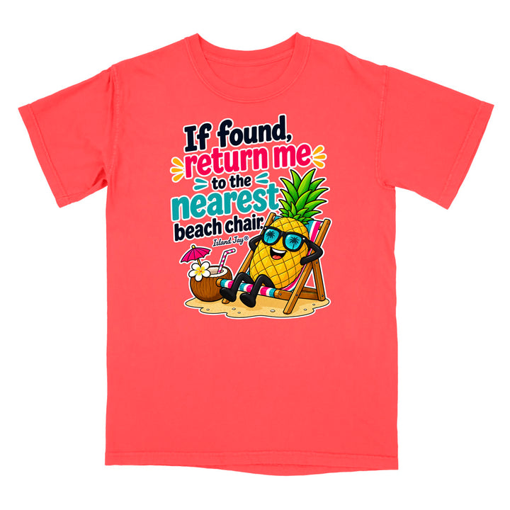 A coral red short-sleeve t-shirt with a colorful graphic on the front. The graphic features a cartoon pineapple wearing sunglasses and relaxing in a beach chair next to a coconut drink, with the text 'If found, return me to the nearest beach chair' and 'Made by: The Pineapple Company'.