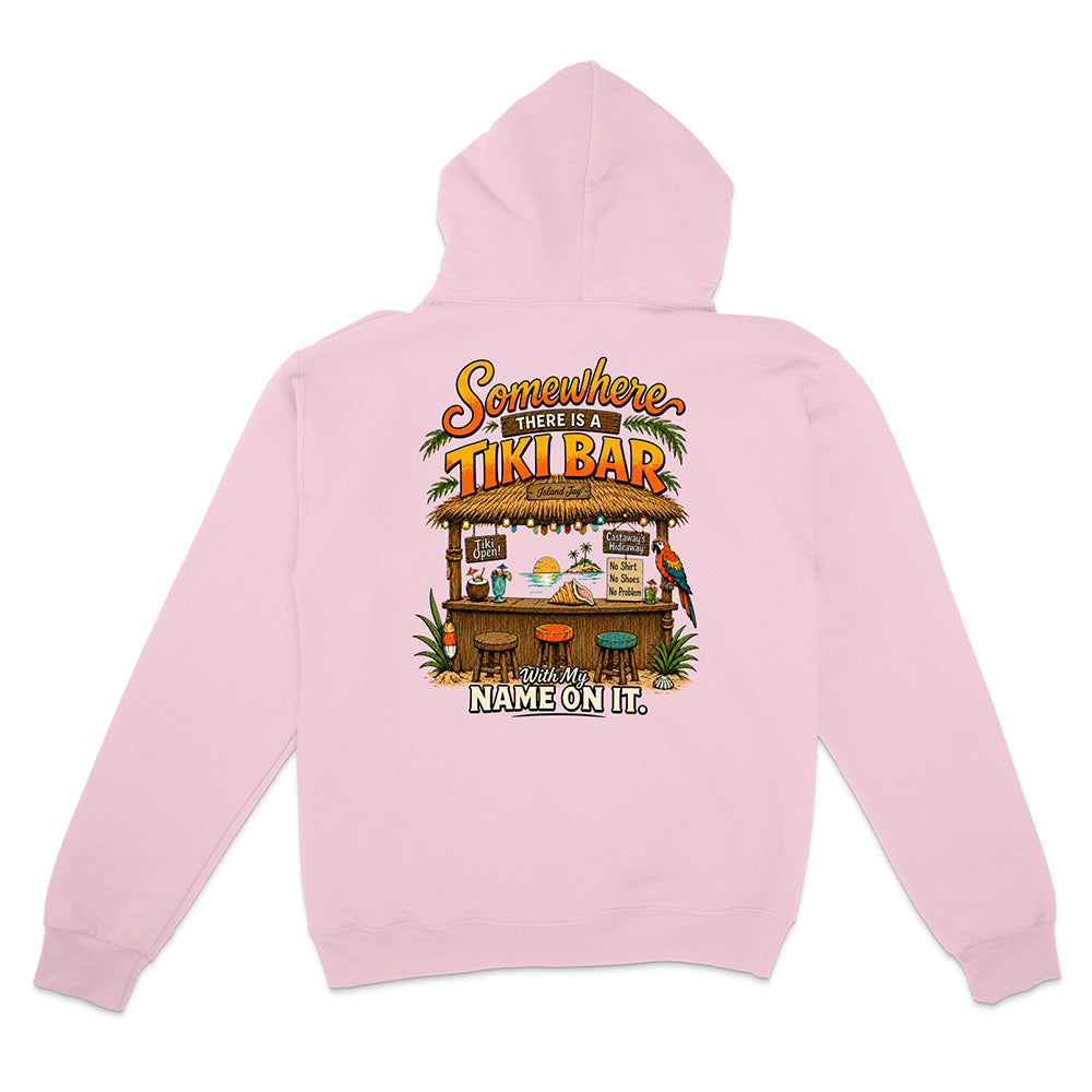A light pink hoodie shown from the back, featuring a colorful graphic print of a tropical 'Tiki Bar' with the text 'Somewhere in between a TIKI BAR' and 'NAME ON IT'.