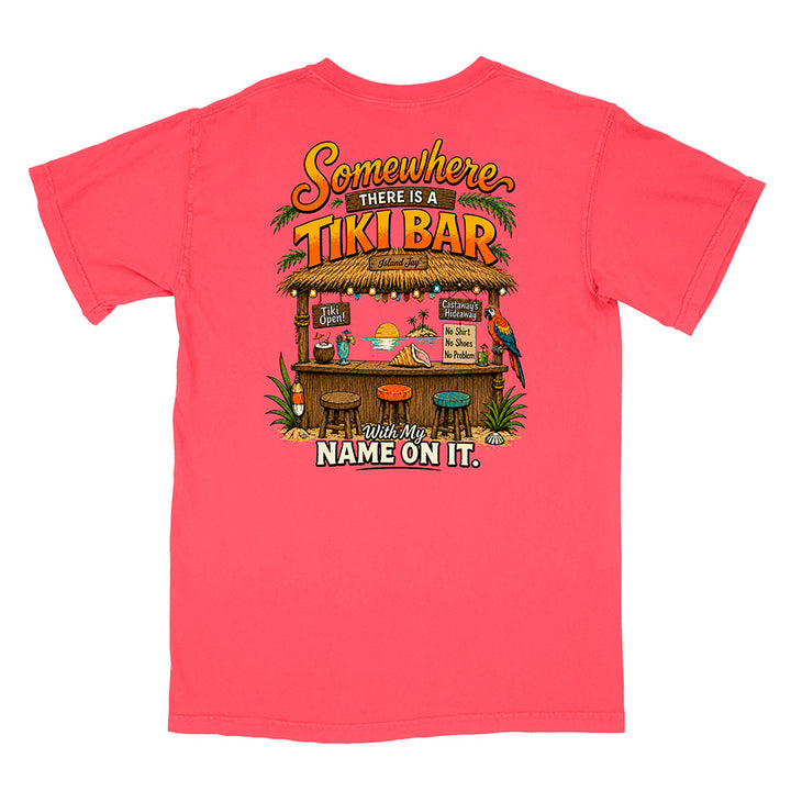 A coral pink short-sleeve t-shirt with a colorful graphic on the back featuring a tropical tiki bar scene and the text 'Somewhere THERE IS A TIKI BAR with My NAME ON IT.'.