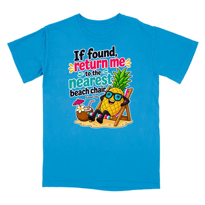 A bright blue short-sleeve t-shirt with a colorful graphic on the front. The graphic features a cartoon pineapple wearing sunglasses and sitting in a beach chair next to a coconut drink, with the text 'If found, return me to the nearest beach chair' in playful multicolor lettering.