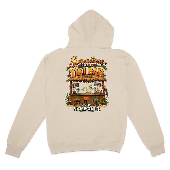 A beige pullover hoodie shown from the back, featuring a colorful graphic print of a tropical 'Tiki Bar' with the text 'Somewhere in between a TIKI BAR' and 'NAME ON IT.'.