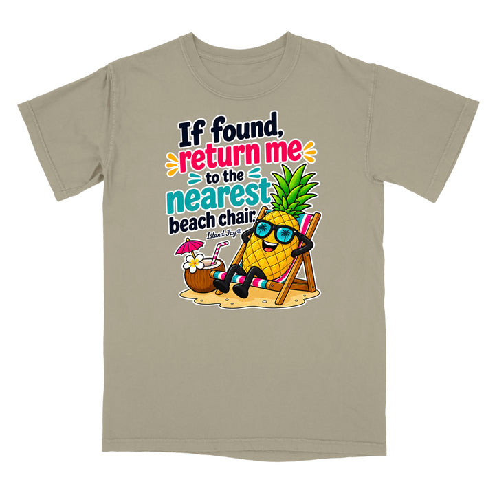 A light taupe crewneck t-shirt featuring a colorful graphic of a pineapple character wearing sunglasses and sitting in a beach chair next to a coconut drink, with the text 'If found, return me to the nearest beach chair' and 'Shark Ray'.