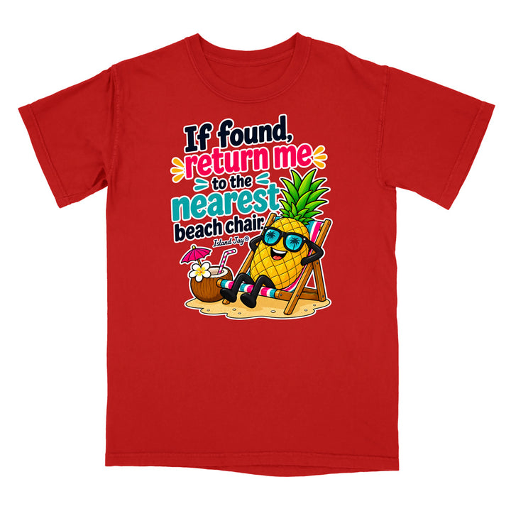 A red short-sleeve crewneck t-shirt featuring a colorful graphic print of a pineapple character wearing sunglasses and sitting in a beach chair next to a coconut drink, with the text 'If found, return me to the nearest beach chair' in playful multicolor lettering.