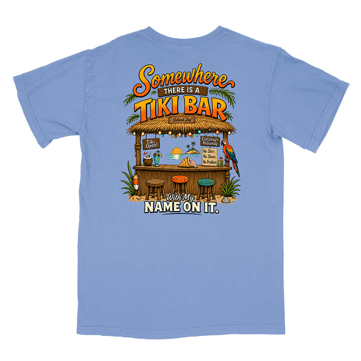 A light blue short-sleeve t-shirt shown flat against a white background, featuring a large graphic print on the back with a tropical tiki bar scene and the text 'Somewhere THERE IS A TIKI BAR With My NAME ON IT.'.