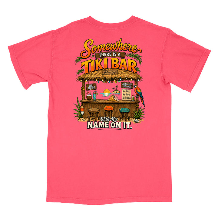 A bright pink short-sleeve t-shirt shown flat against a white background, featuring a colorful graphic on the front with the text 'Somewhere THERE IS A TIKI BAR' and 'Make My NAME ON IT.' above and below a tropical tiki bar illustration.