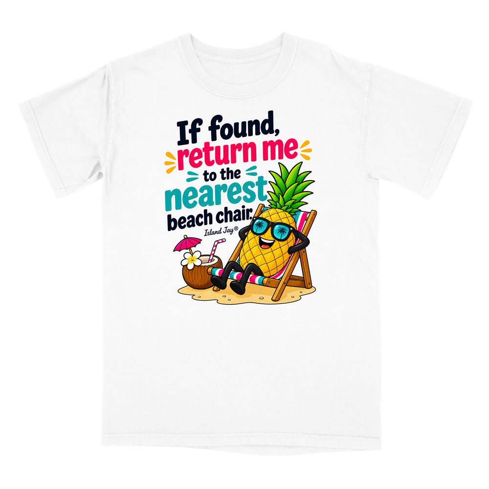 A white short-sleeve t-shirt with a colorful graphic print on the front. The graphic features a cartoon pineapple wearing sunglasses and reclining in a beach chair next to a coconut drink with a pink umbrella, with the text 'If found, return me to the nearest beach chair.