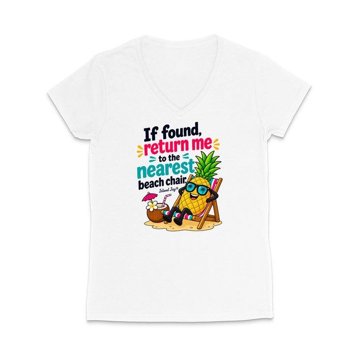 A white women's V-neck t-shirt with a colorful graphic print on the front. The graphic features a cartoon pineapple wearing sunglasses and sitting in a beach chair next to a coconut drink, with the text 'If found, return me to the nearest beach chair' in black, pink, and teal lettering.