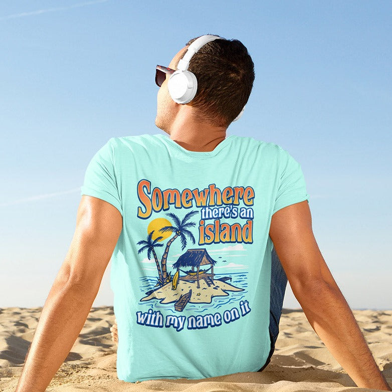 A man relaxing at the beach and siting in the sand. He is wearing Island Jay's Tropical Men's Cotton T-Shirt with Somewhere The Is An Island written on it. Island Reef Green