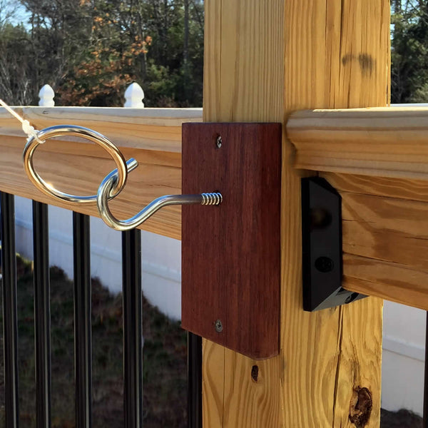 A solid mahogany hook and eye latch, inspired by those found in the classic Ring Toss Pro Quick Shot game, is attached to a wooden post to secure a horizontal rail. Against a background of black vertical bars and an outdoor scene with trees and fencing, this piece showcases timeless craftsmanship and quality—proudly made in the USA.
